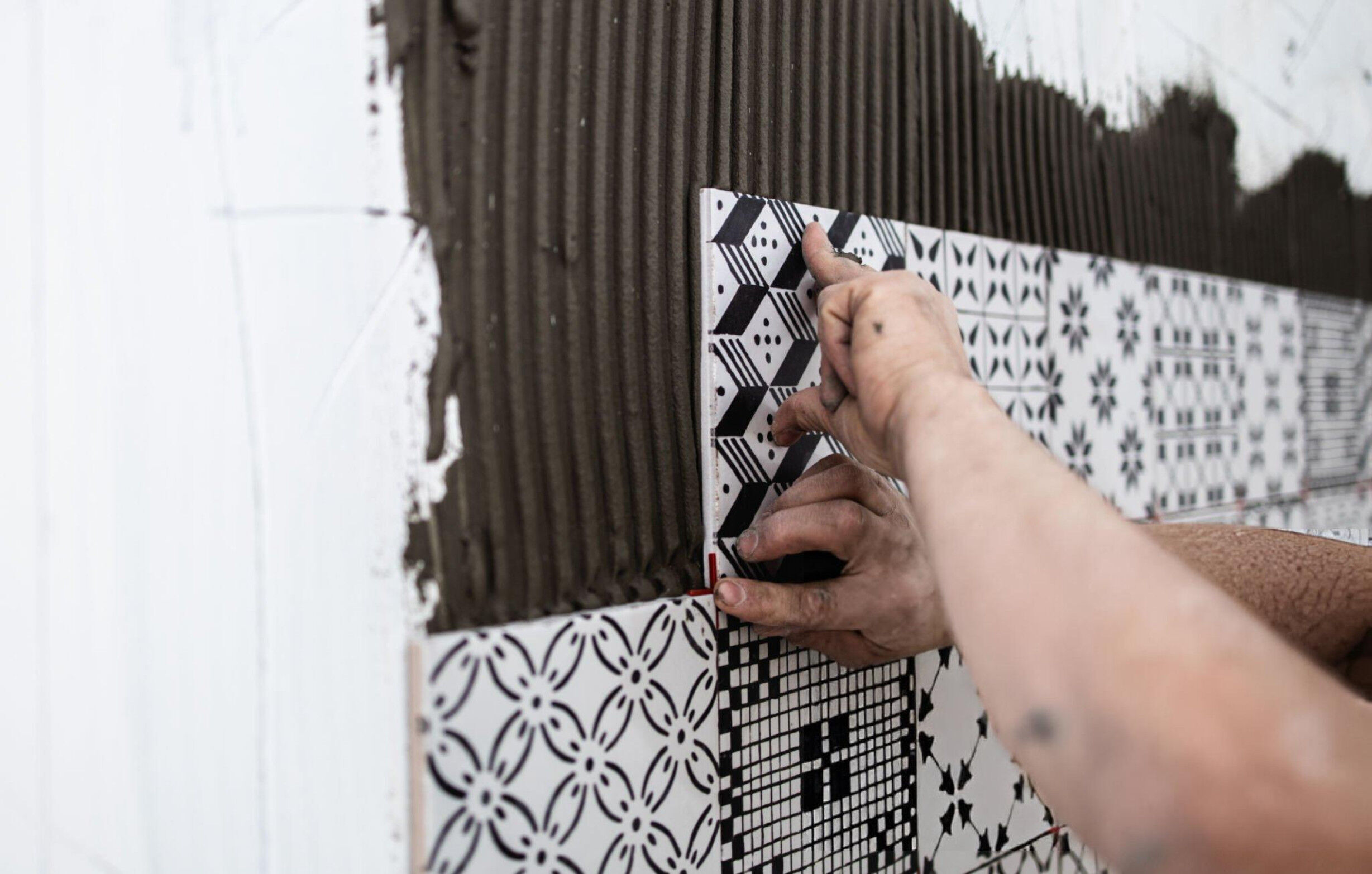 Hands installing patterned black and white tiles on a bathroom wall with adhesive