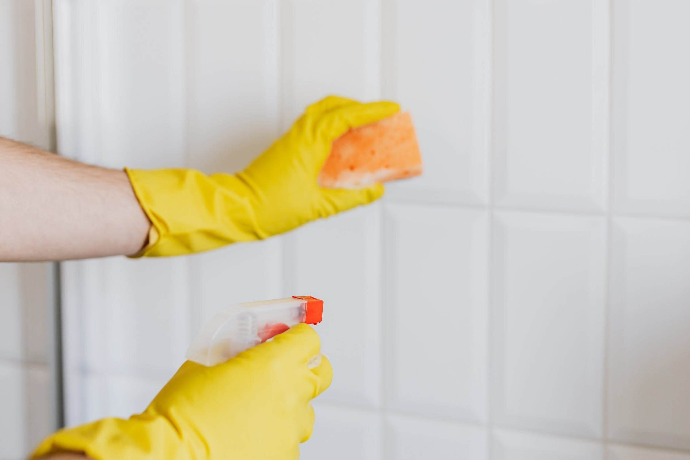 Person in yellow gloves cleaning white tiles with a sponge and spray bottle