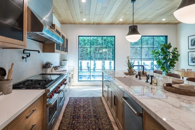 Modern kitchen with marble island, wooden cabinets, and large windows providing natural light