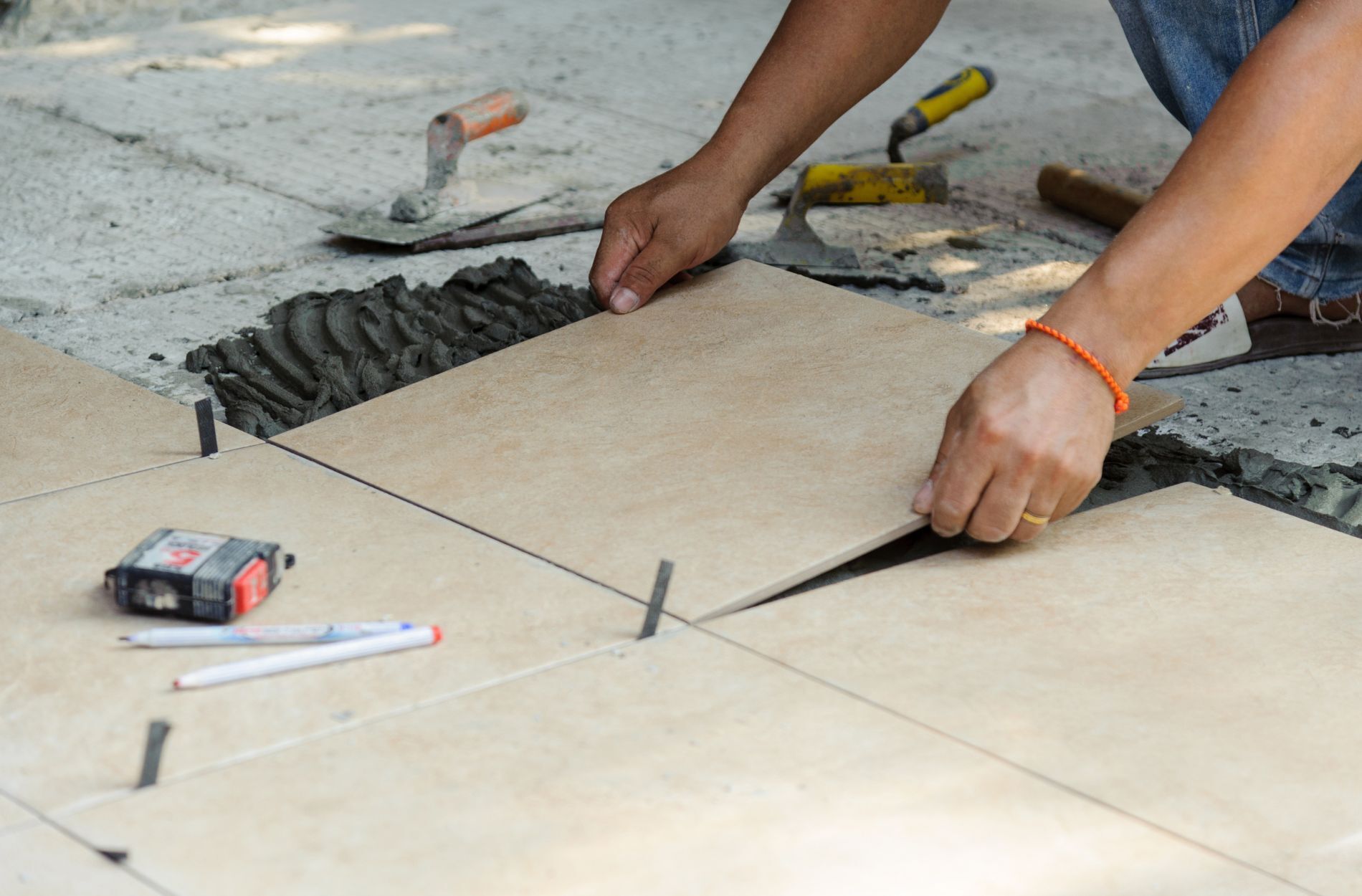 Person installing large beige tiles outdoors with tools and adhesive visible