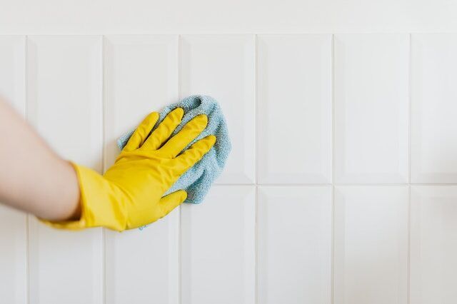 Person in yellow glove cleaning white tiled wall with blue cloth