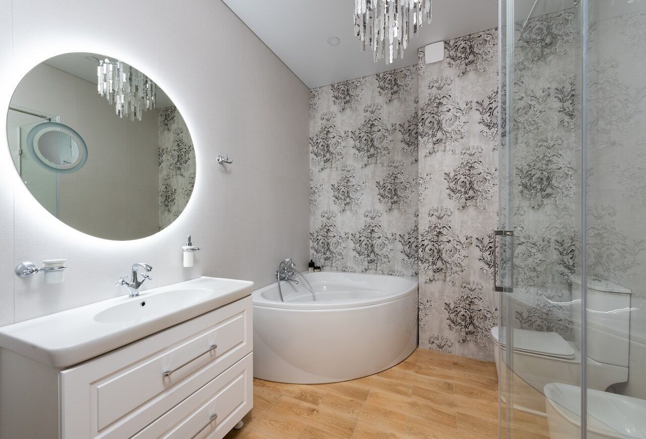 Modern small bathroom with illuminated mirror, white vanity, corner bathtub, and patterned wallpaper