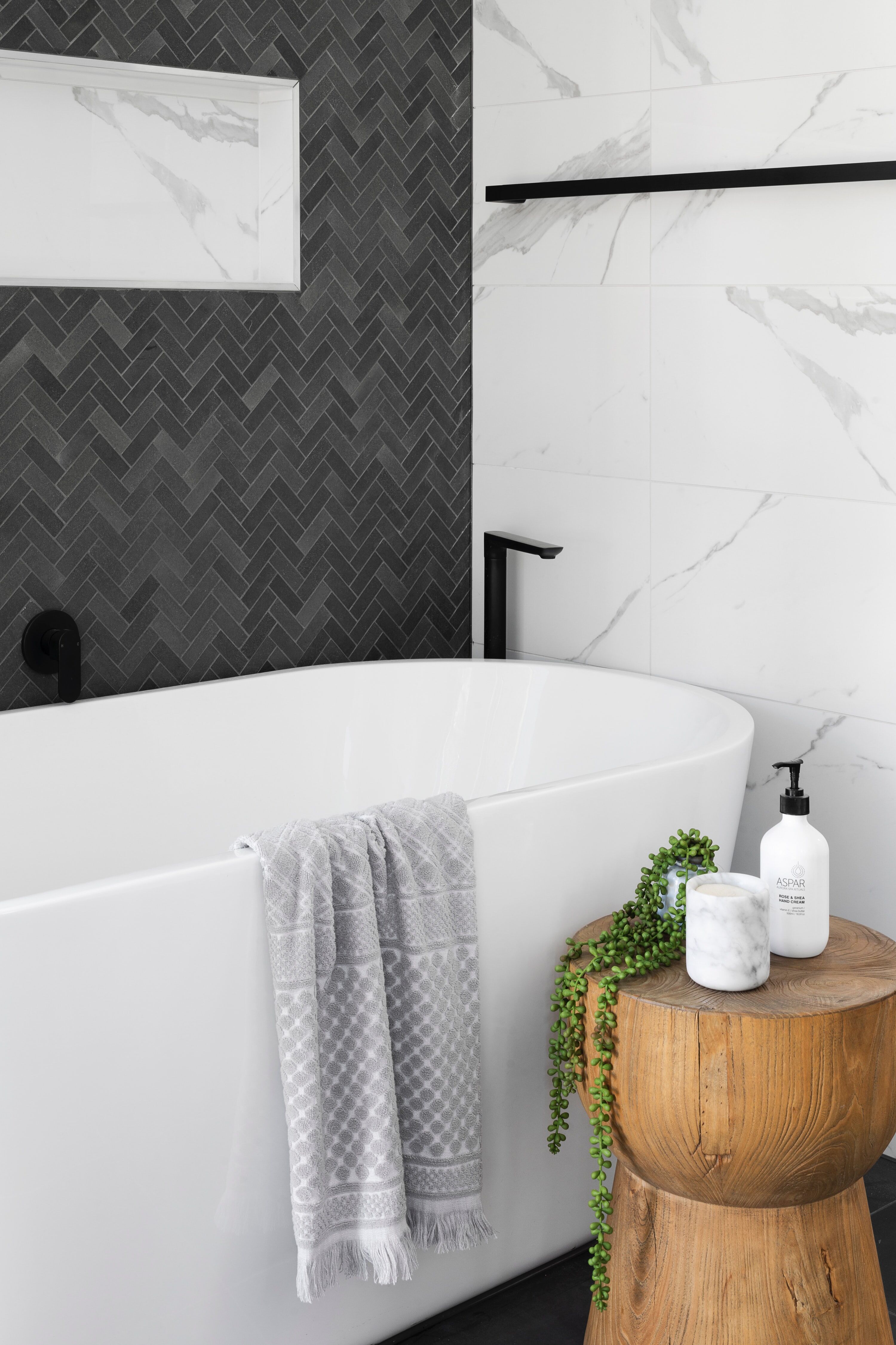 White freestanding bathtub with gray towel, dark herringbone tile wall, and wooden stool with plant and toiletries