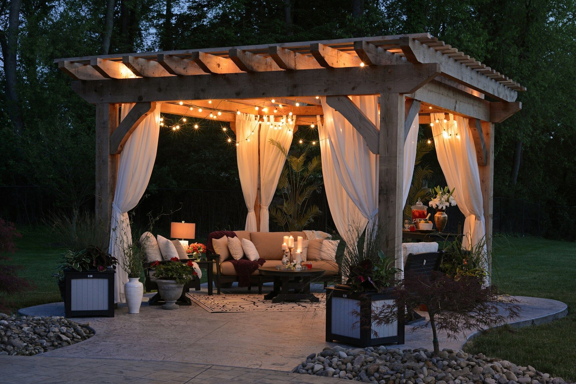 Outdoor patio with wooden pergola, string lights, and white curtains, surrounded by plants