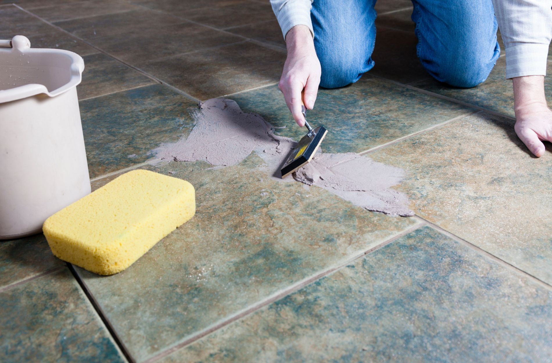 Person applying grout to a tiled floor with a grout float, sponge, and bucket nearby