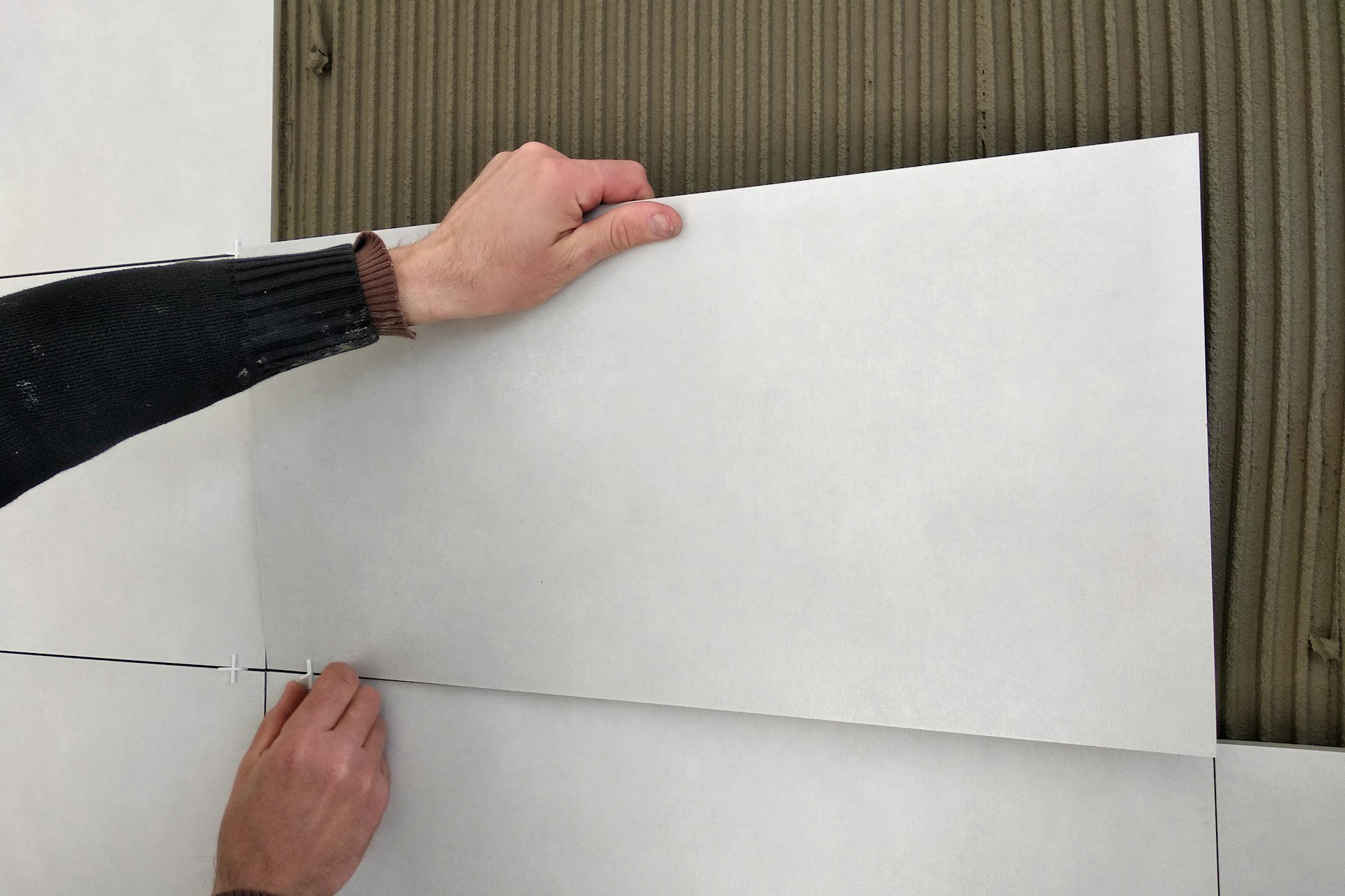 Hands installing large white tiles on a wall with spacers and adhesive