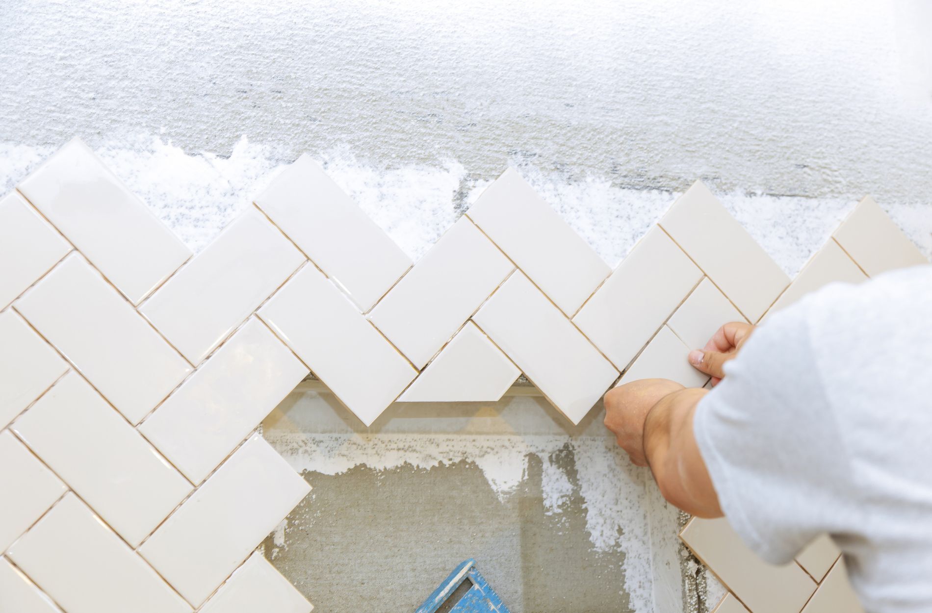Person installing beige ceramic tiles in a herringbone pattern on a wall with visible adhesive